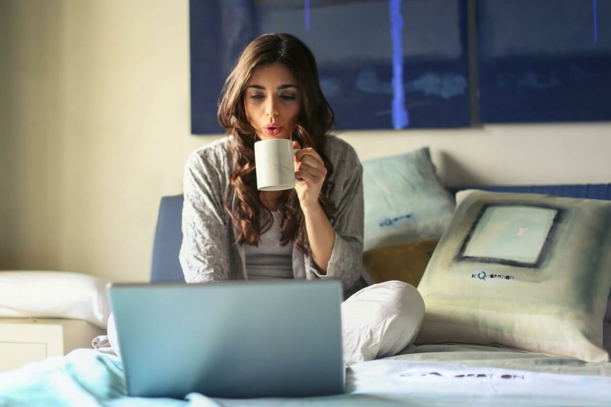 Woman sitting in bed drinking coffee and working on a laptop. Photo credit Andrea Piacquadio