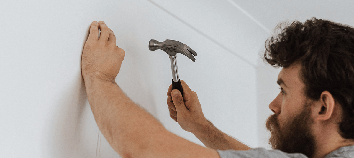 Man using a hammer to pound a picture hanger into a wall. Image credit: Anete Lusina